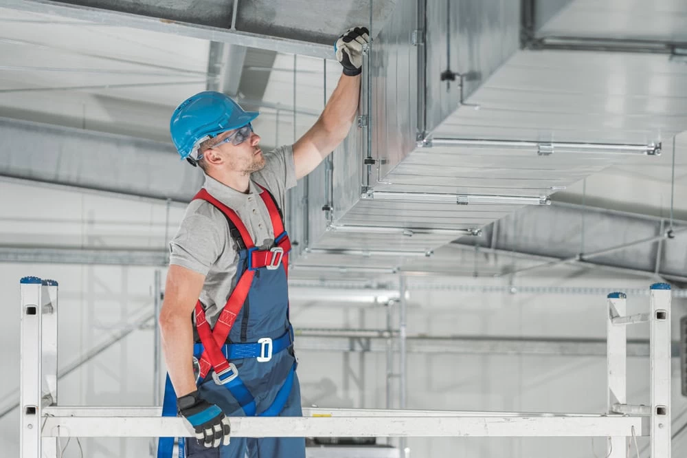 A construction worker wearing a blue helmet, safety glasses, gloves, and a red harness works on metal ductwork while standing on scaffolding indoors. He is reaching up to make adjustments to the ducting system in a spacious, industrial setting, showcasing skills akin to those used in Residential Duct Cleaning Sacramento.