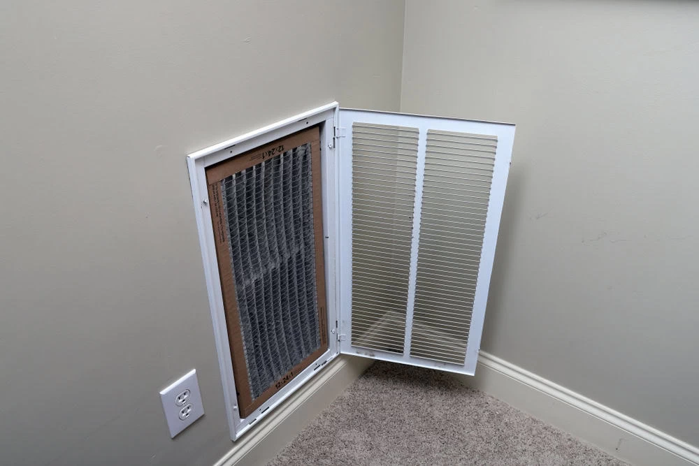 An open wall vent with a visible air filter located low on an interior wall, near the floor. The vent cover, attached to the wall with hinges, is open to the right. A power outlet is positioned just below the vent. For optimal performance and cleanliness, consider Air Duct Cleaning Sacramento services.