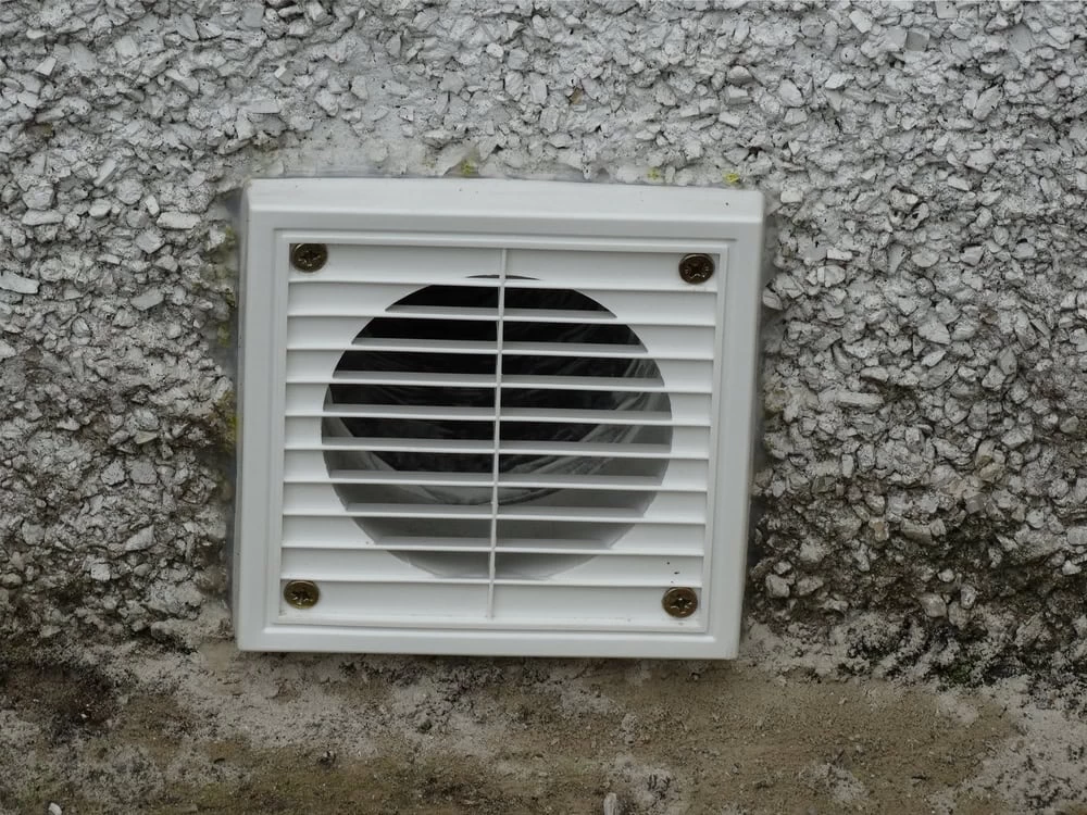 Close-up of a white vent mounted on a textured, gray exterior wall. The vent, likely part of an exhaust duct cleaning Sacramento system, features horizontal slats covering a round opening, secured by four screws. Moss is visible at the base of the wall.