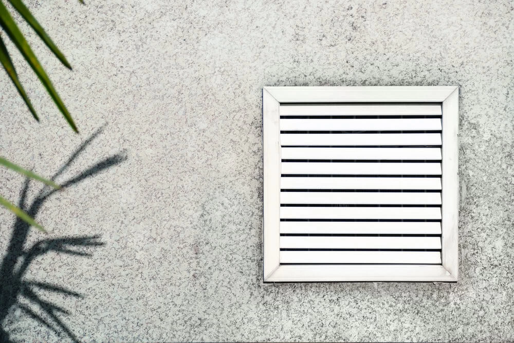 A white, square ventilation grill with horizontal slats is mounted on a speckled gray wall. The shadow of a plant with long, slender leaves is cast on the left side of the wall. For optimal performance and air quality, consider scheduling a professional Ventilation Duct Cleaning Sacramento service.