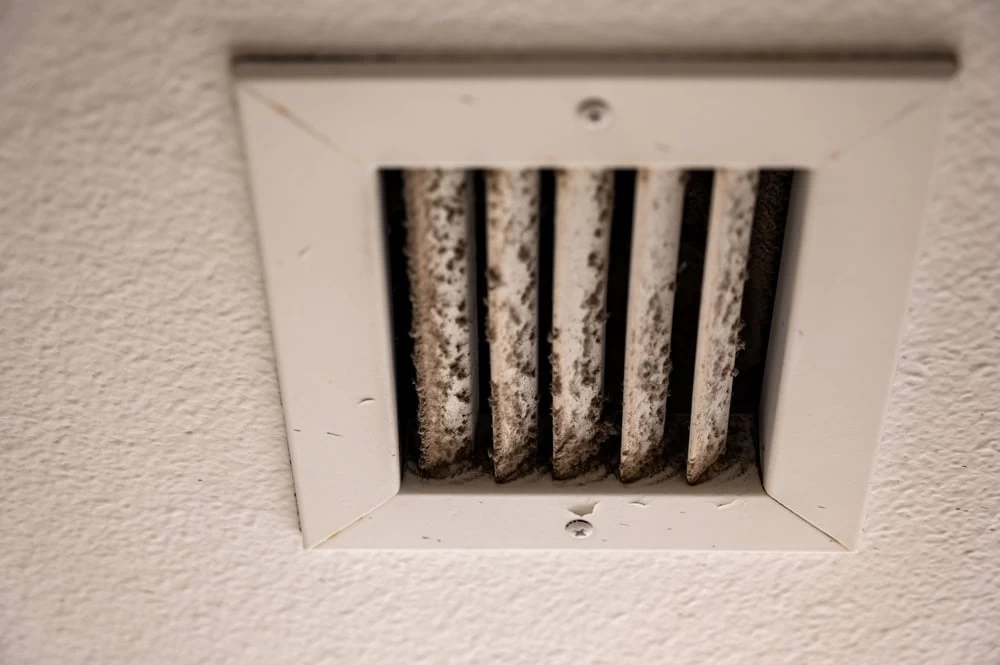 A close-up image of a white ceiling air vent with several vertical slats. The vent is dirty, with visible dust and grime accumulated on the slats, highlighting the need for Residential Duct Cleaning Sacramento services. The surrounding white ceiling also shows some minor dirt and wear.