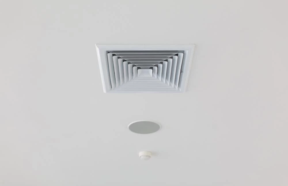 Close-up of a white ceiling featuring a large square ventilation grill, a small round smoke detector, and a circular recessed light fixture. The smooth, white ceiling showcases evenly spaced devices, ideal for those considering Residential Duct Cleaning Sacramento to maintain optimal air quality.