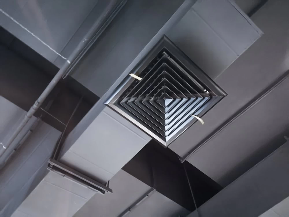 Close-up view of an industrial-style ceiling vent with geometric, triangular layers in a modern building. The ceiling is painted grey with visible structural elements and pipework, a testament to the thorough HVAC Cleaning Sacramento ensures for peak efficiency and aesthetics.