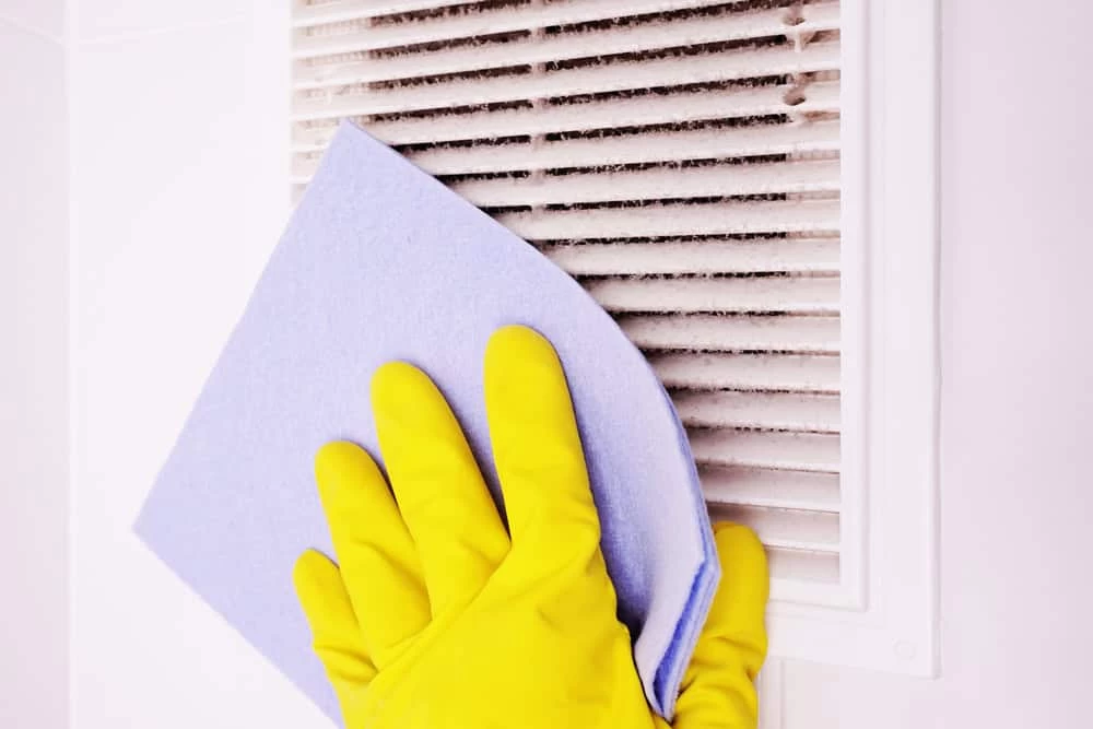 A person wearing bright yellow rubber gloves uses a blue cloth to clean a dirty white air vent, showcasing the meticulous care typical of Residential Duct Cleaning Sacramento. The vent, attached to a wall, has visible dust and dirt accumulated on it.