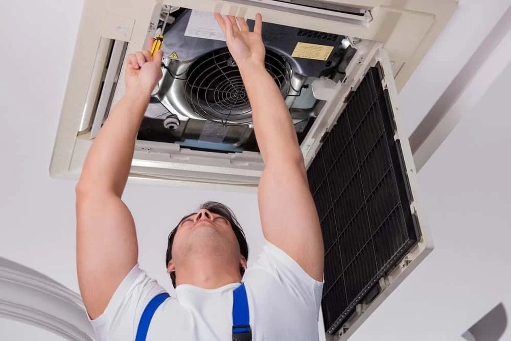 A person wearing a white shirt and blue overalls is fixing an overhead air conditioning unit, part of an HVAC Cleaning Sacramento service. They are using tools to work on the unit's internal components, and the open cover reveals the fan and other mechanical parts.