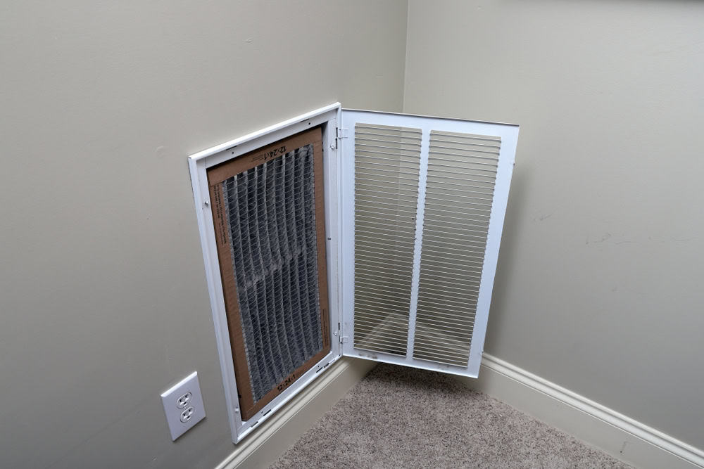 An open wall vent with a visible air filter located low on an interior wall, near the floor. The vent cover, attached to the wall with hinges, is open to the right. A power outlet is positioned just below the vent. For optimal performance and cleanliness, consider Air Duct Cleaning Sacramento services.