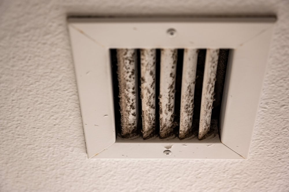 A close-up image of a white ceiling air vent with several vertical slats. The vent is dirty, with visible dust and grime accumulated on the slats, highlighting the need for Residential Duct Cleaning Sacramento services. The surrounding white ceiling also shows some minor dirt and wear.