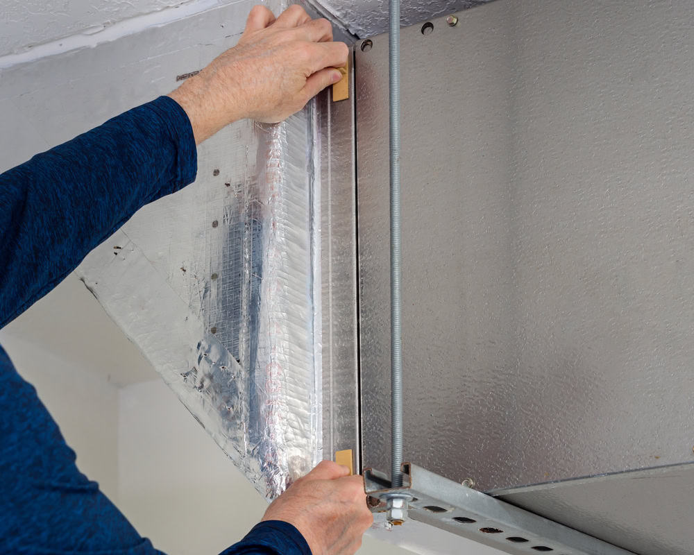 Close-up of a person securing metal ductwork with insulation material. They are using their hands to fasten the insulation around the duct, ensuring a tight fit. The person is wearing a blue long-sleeve shirt. Ideal for services like Residential Duct Cleaning Sacramento, the image focuses on the hands and ductwork.