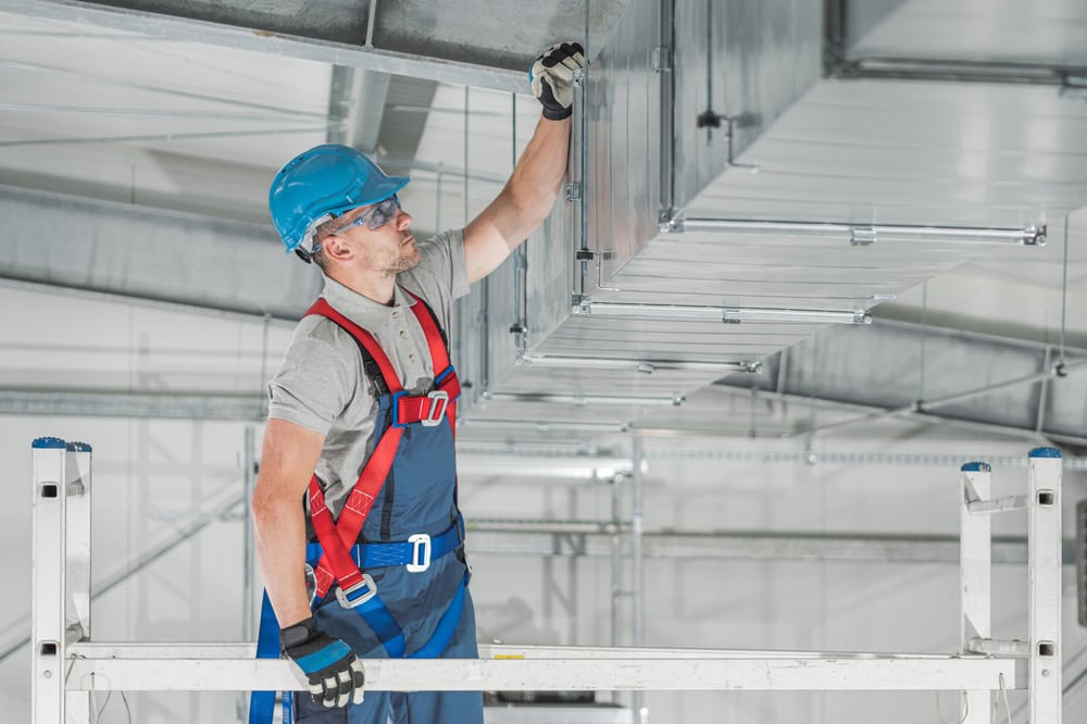A construction worker wearing a blue helmet, safety glasses, gloves, and a red harness works on metal ductwork while standing on scaffolding indoors. He is reaching up to make adjustments to the ducting system in a spacious, industrial setting, showcasing skills akin to those used in Residential Duct Cleaning Sacramento.