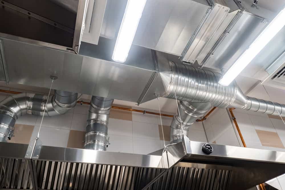 A view of a modern commercial kitchen ceiling with shiny metal ventilation ducts, stainless steel exhaust hoods, and bright fluorescent lights. The ducts and piping are neatly arranged, creating an industrial yet clean appearance—ideal for those considering Air Duct Cleaning Sacramento services.