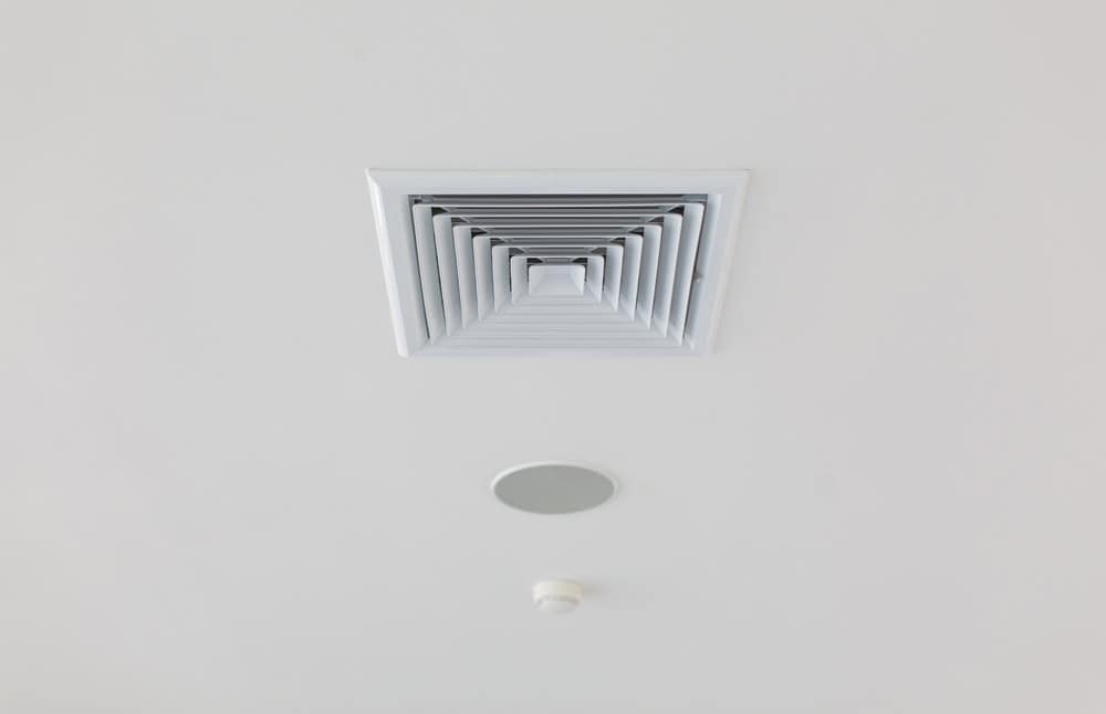 Close-up of a white ceiling featuring a large square ventilation grill, a small round smoke detector, and a circular recessed light fixture. The smooth, white ceiling showcases evenly spaced devices, ideal for those considering Residential Duct Cleaning Sacramento to maintain optimal air quality.
