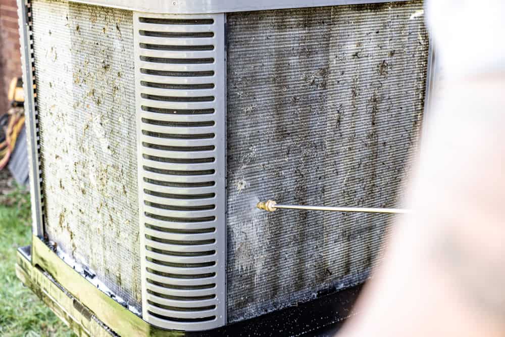 A person is pressure washing the dirty exterior of an outdoor air conditioning unit. The unit is stained with debris and grime, highlighting the need for maintenance—a task as essential as HVAC Cleaning Sacramento. The pressure washer's water nozzle is close to the surface of the unit.