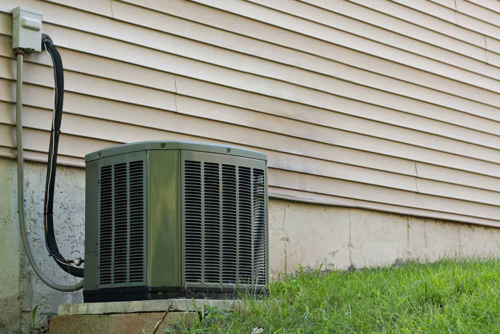 A green outdoor air conditioning unit sits on a concrete pad next to the exterior wall of a house with light-colored siding. The unit's electrical wiring is visible, attached to the side of the house. Nearby, Residential Duct Cleaning Sacramento ensures optimal air quality in this grassy area.