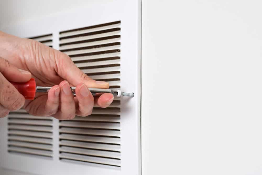 Close-up of a person using a red-handled screwdriver to tighten a screw on a white wall vent cover. The person's hands are focused on securing the screw. Ensuring proper maintenance like this can be part of thorough Air Duct Cleaning Sacramento services. The vent cover has horizontal slats for air passage.