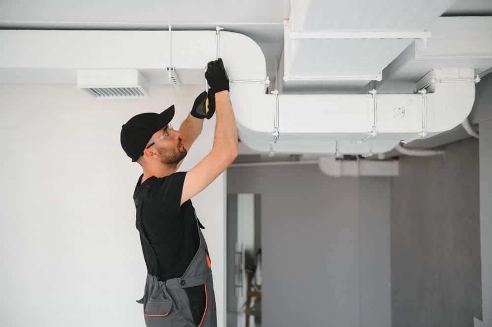A person in a black cap, black shirt, and gray overalls is using a screwdriver to fix or install a white duct system on the ceiling. The individual, likely involved in HVAC Cleaning Sacramento, is wearing black gloves and working in a modern, light-colored room.