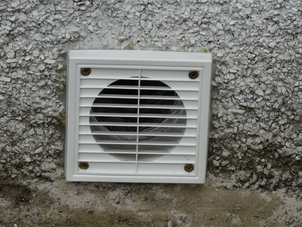 Close-up of a white vent mounted on a textured, gray exterior wall. The vent, likely part of an exhaust duct cleaning Sacramento system, features horizontal slats covering a round opening, secured by four screws. Moss is visible at the base of the wall.