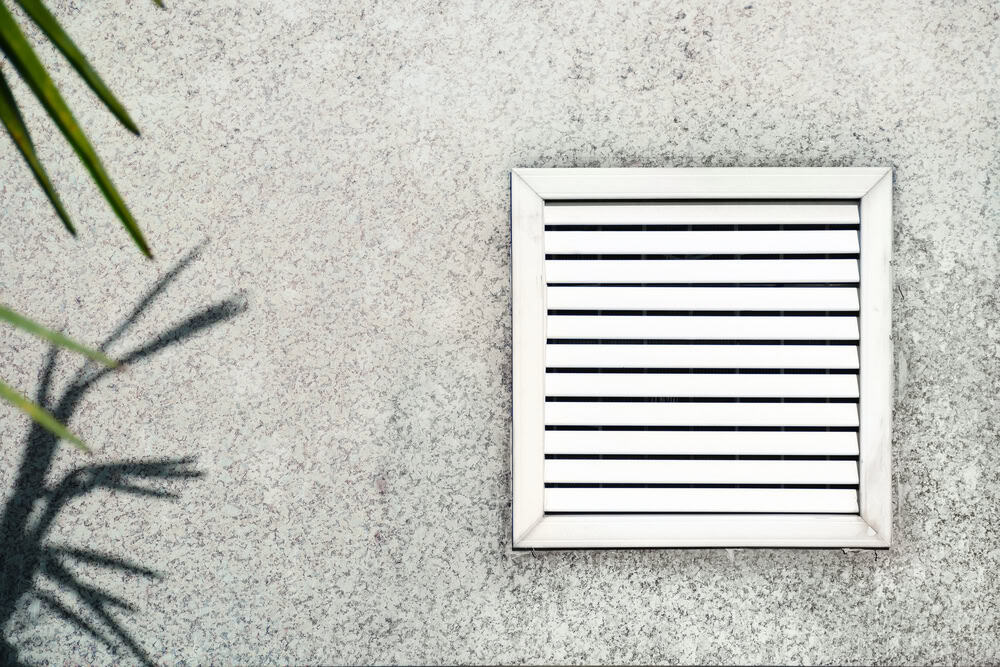 A white, square ventilation grill with horizontal slats is mounted on a speckled gray wall. The shadow of a plant with long, slender leaves is cast on the left side of the wall. For optimal performance and air quality, consider scheduling a professional Ventilation Duct Cleaning Sacramento service.