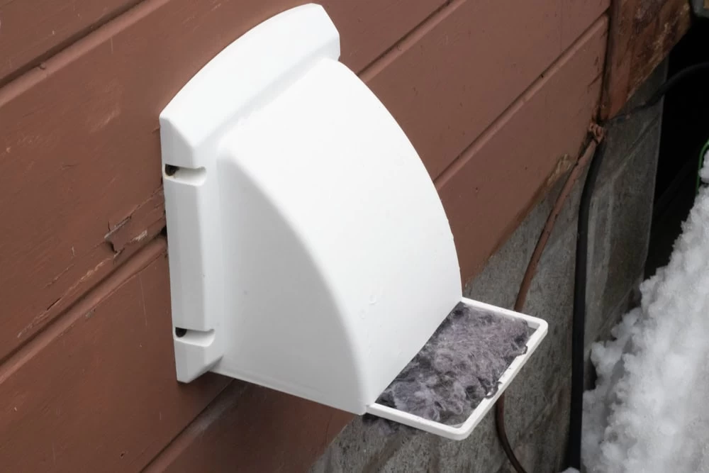 A white exterior dryer vent cover attached to the side of a brown wooden house. The vent cover is open, revealing a removable lint trap filled with gray lint. As snow blankets the ground near the foundation, it's clear that Dryer Vent Cleaning Sacramento services might be needed soon.