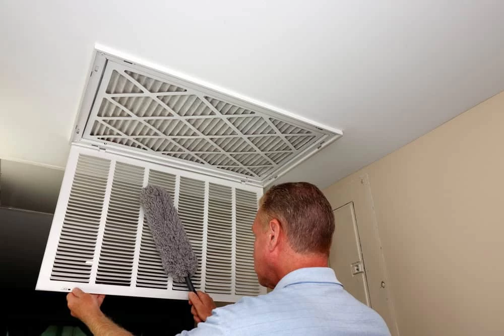 A person wearing a light blue shirt is using a duster to clean an air vent located on the ceiling. The vent cover is open, revealing a filter. The surrounding beige and white walls emphasize the importance of Air Duct Cleaning Sacramento for maintaining indoor air quality.