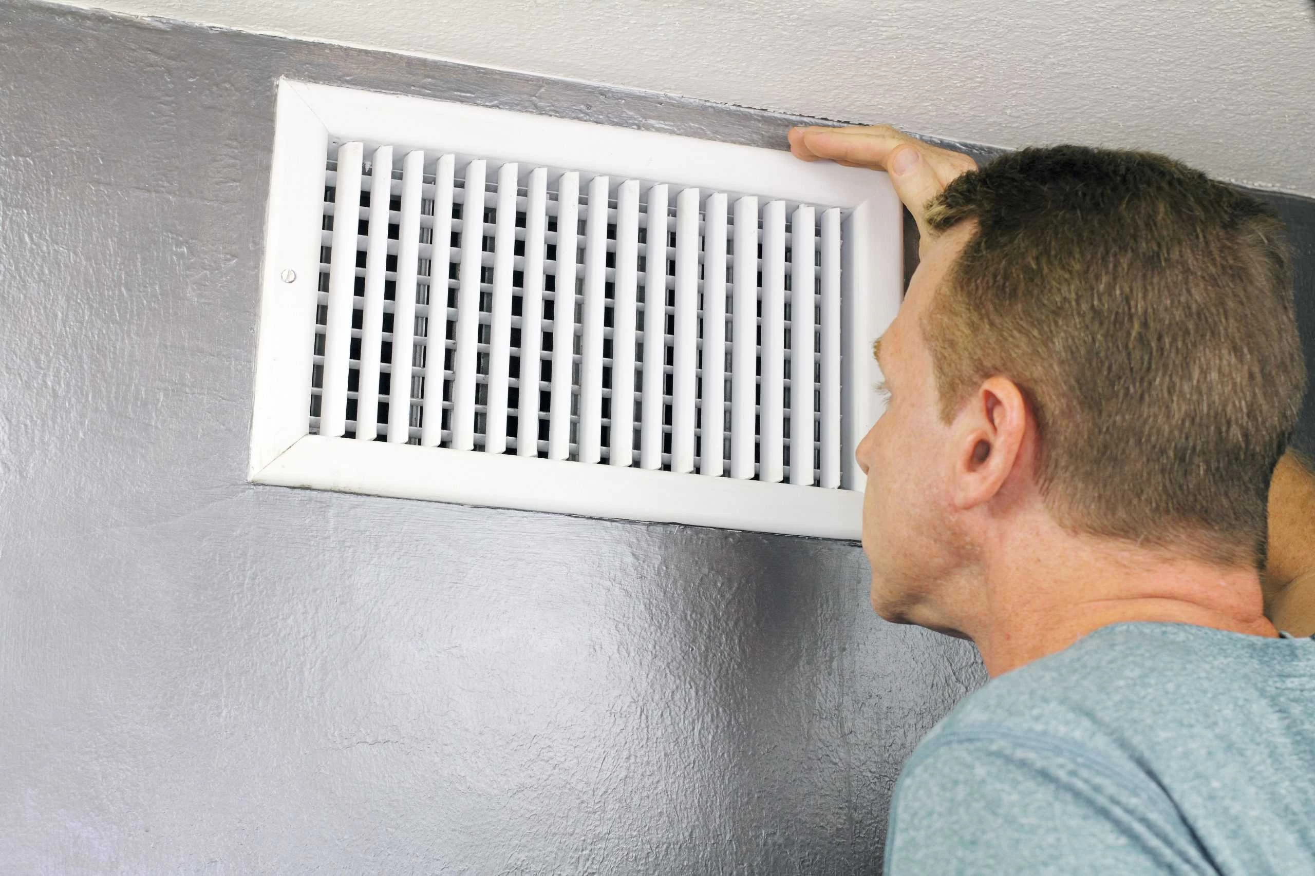 A person inspecting a rectangular white air vent on a gray wall, possibly checking for cleanliness or proper function. The individual, dressed in a light blue shirt with short hair, appears to be ensuring the effectiveness of residential duct cleaning in Sacramento.