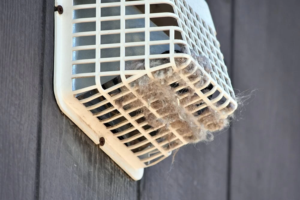 A white outdoor dryer vent cover, clogged with lint, is attached to a dark gray wooden wall. The thick buildup blocks the vent's openings, highlighting the need for professional HVAC Cleaning Sacramento services.
