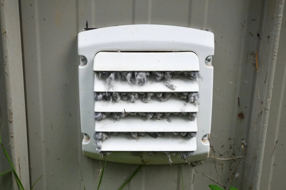 A white dryer vent clogged with lint is attached to a beige exterior wall, surrounded by grass. The partially open vent flaps reveal accumulated lint inside, highlighting the need for professional services like HVAC Cleaning Sacramento.