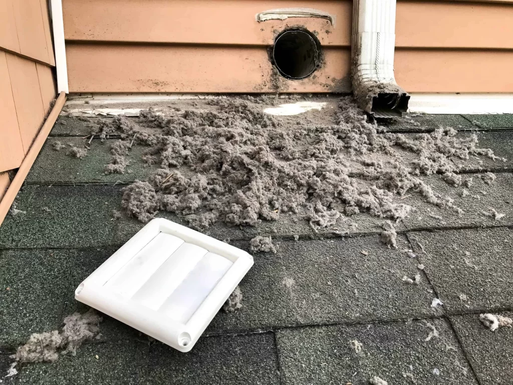 A broken dryer vent cover lies on the roof with a large amount of lint scattered around it, highlighting the need for Exhaust Duct Cleaning Sacramento. The exposed vent opening and part of a drainpipe are visible nearby. Green shingles cover the roof, complementing the light brown house siding.