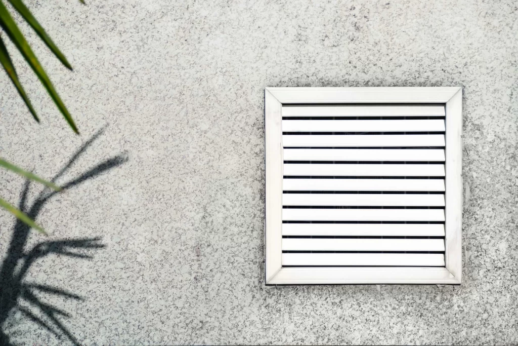 A square ventilation grate with horizontal slats is installed on a textured concrete wall. The shadow of a plant with spiky leaves is cast on the wall to the left of the grate, highlighting areas that may benefit from Residential Duct Cleaning Sacramento for optimal airflow.