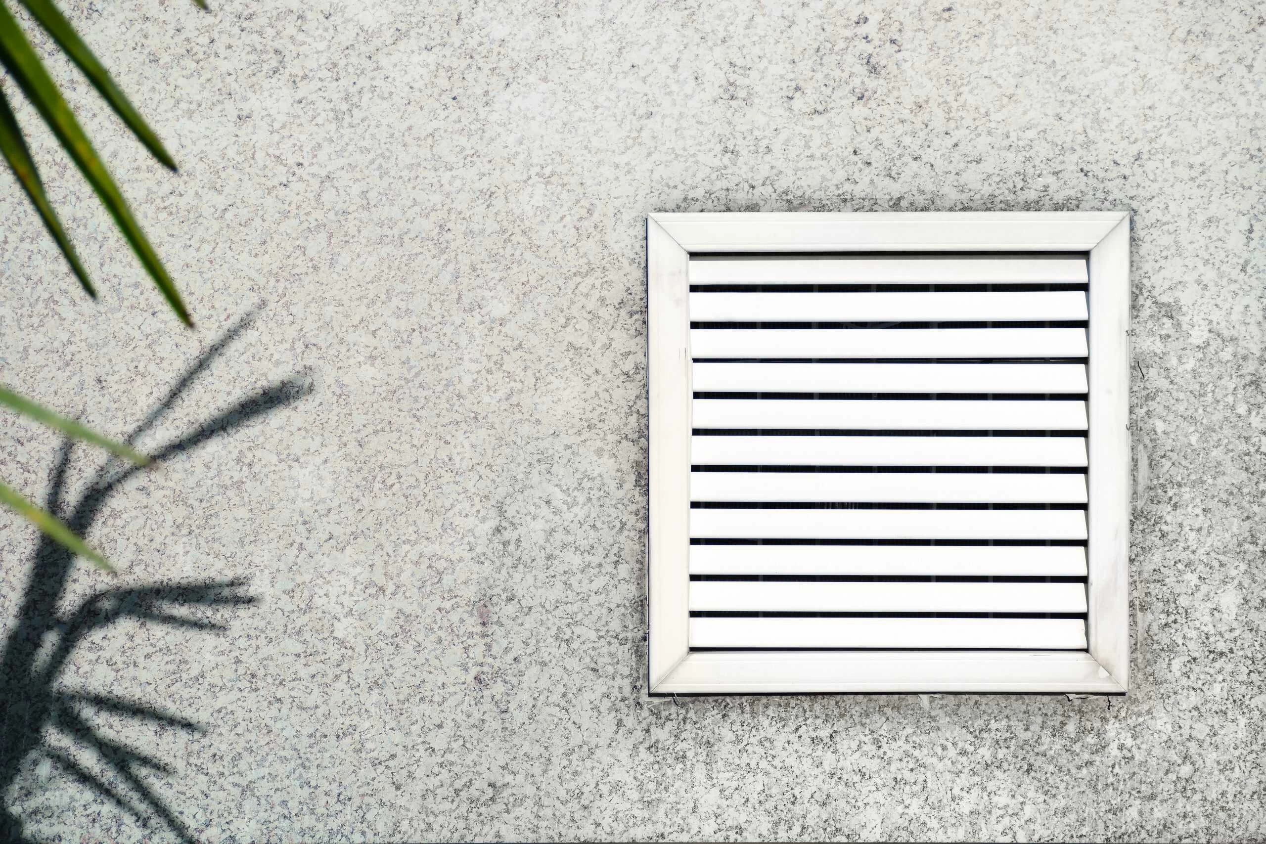 A square ventilation grate with horizontal slats is installed on a textured concrete wall. The shadow of a plant with spiky leaves is cast on the wall to the left of the grate, highlighting areas that may benefit from Residential Duct Cleaning Sacramento for optimal airflow.