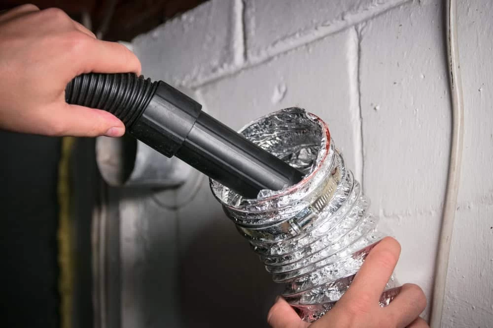 Two hands are shown cleaning the inside of a silver, flexible duct with a black vacuum hose. The duct appears to be part of a venting system attached to a white wall, possibly in a basement or utility area. This scene highlights professional Dryer Vent Cleaning Sacramento services in action.