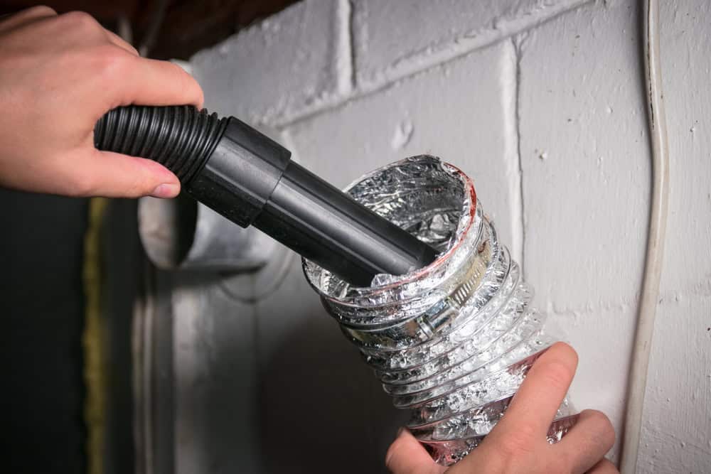 Two hands are shown cleaning the inside of a silver, flexible duct with a black vacuum hose. The duct appears to be part of a venting system attached to a white wall, possibly in a basement or utility area. This scene highlights professional Dryer Vent Cleaning Sacramento services in action.