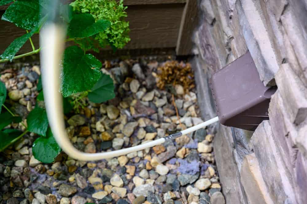 A hose is connected to a brown box mounted on a stone wall, part of an Air Duct Cleaning Sacramento setup. The ground beneath is covered with small pebbles, and green plants are seen nearby.