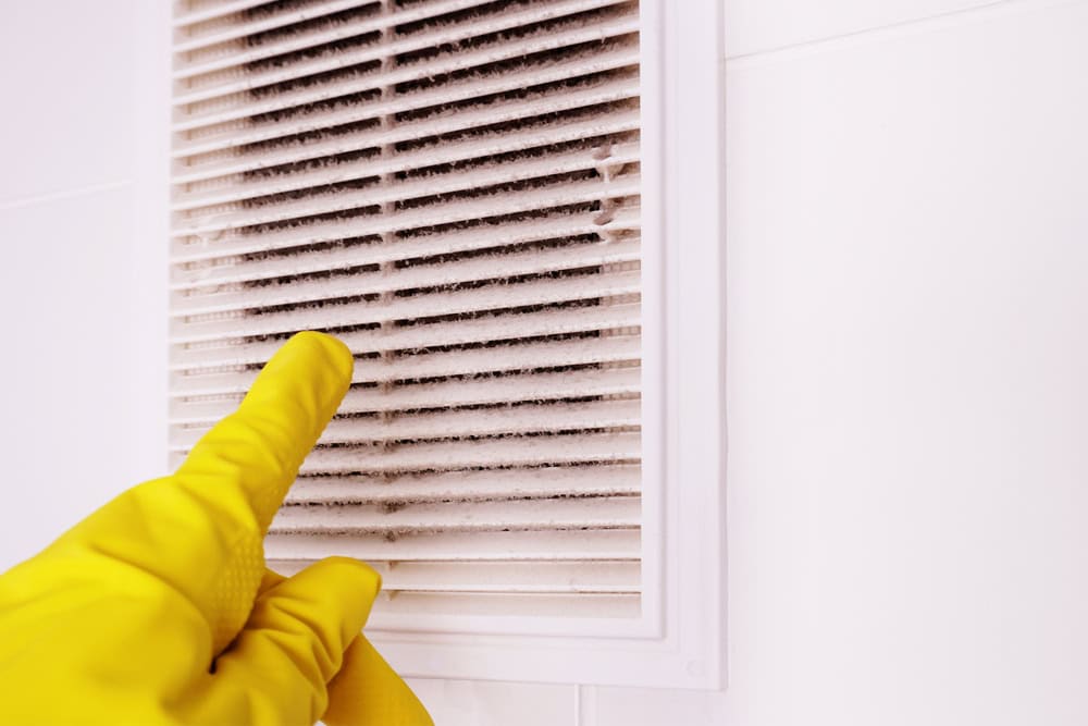 A person wearing yellow rubber gloves points to a dirty, dusty air vent on a white wall, emphasizing the need for Air Duct Cleaning Sacramento.