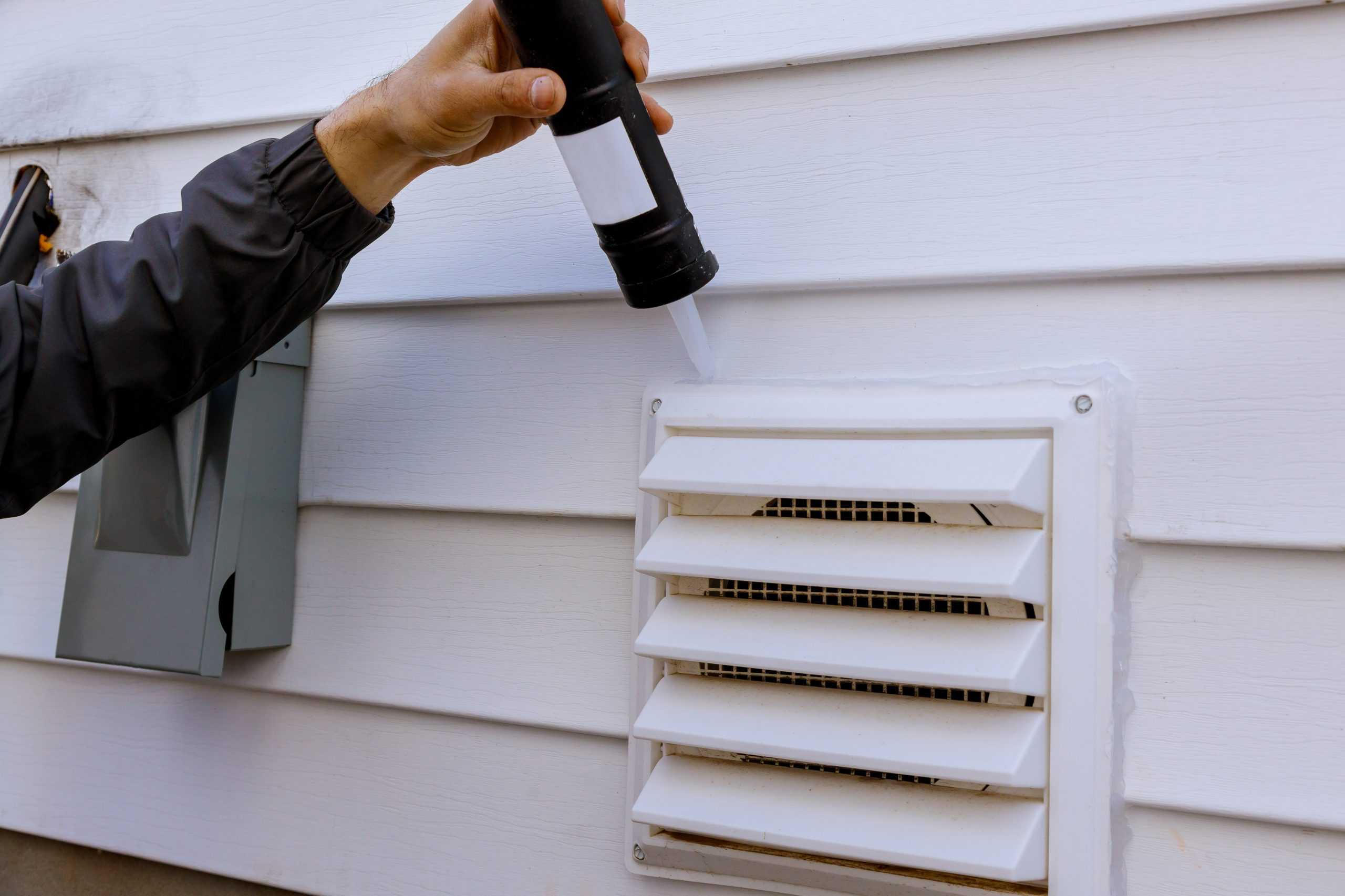 A person applies caulk around a white exterior vent cover on a house, sealing gaps to ensure it is weatherproof. The person, possibly prepping for Residential Duct Cleaning Sacramento, is holding a caulking gun, and part of their sleeve is visible.