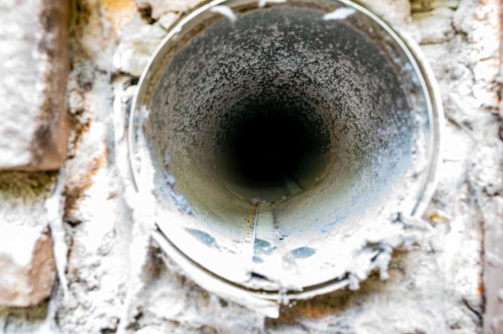 Close-up of the opening of an old and rusted metal pipe mounted in a concrete wall, extending into the darkness. The surrounding area of the pipe is weathered and covered with patches of corrosion and grime. Scenes like this highlight the importance of Residential Duct Cleaning Sacramento for better air quality.