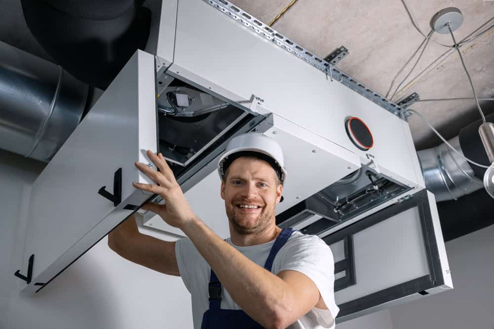 A maintenance worker, wearing a white hard hat and blue overalls, is smiling as he services a piece of HVAC equipment attached to a ceiling. He has opened the front panel of the unit and is looking directly at the camera, exemplifying expert Residential Duct Cleaning Sacramento services.