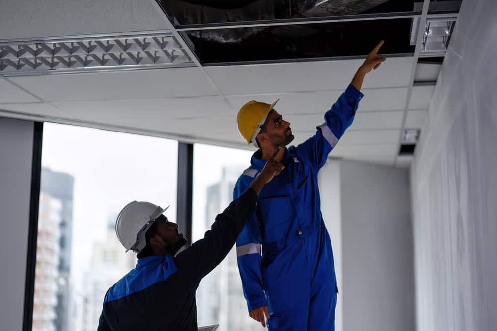Two construction workers in blue coveralls and hard hats inspect the ceiling of an office building. One worker points upwards while the other holds a tablet, both focusing on the ceiling area, possibly checking for issues or conducting maintenance related to Air Duct Cleaning Sacramento.