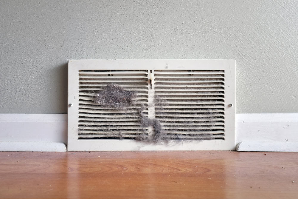 A dusty and dirty white vent cover on a light gray wall, situated just above a hardwood floor. The vent is partially clogged with accumulated dust and debris, particularly in the center of the grille. It's evident that an HVAC Cleaning Sacramento service could restore its efficiency.