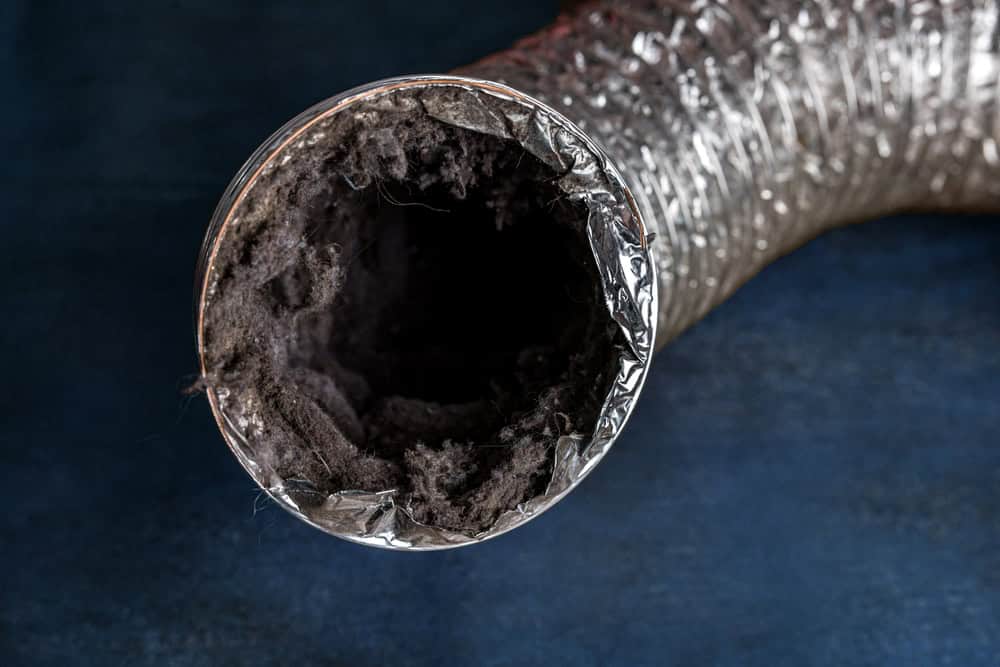 Close-up view of the opening of a flexible, metallic dryer vent hose with visible lint buildup inside. The background is a dark blue surface. Schedule your Dryer Vent Cleaning Sacramento to ensure your home remains safe and efficient.