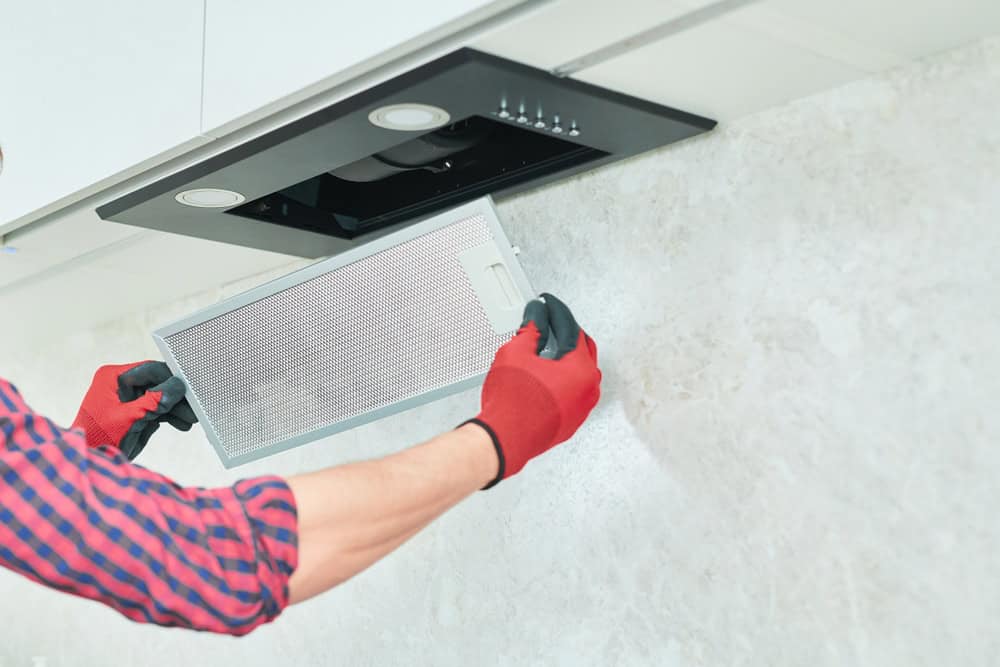 A person with red gloves is removing a mesh filter from a black kitchen range hood installed under a cabinet. The wall behind is light beige, and the individual wears a red and blue checkered shirt, with only their arms visible—a perfect example of expert Air Duct Cleaning Sacramento at work.