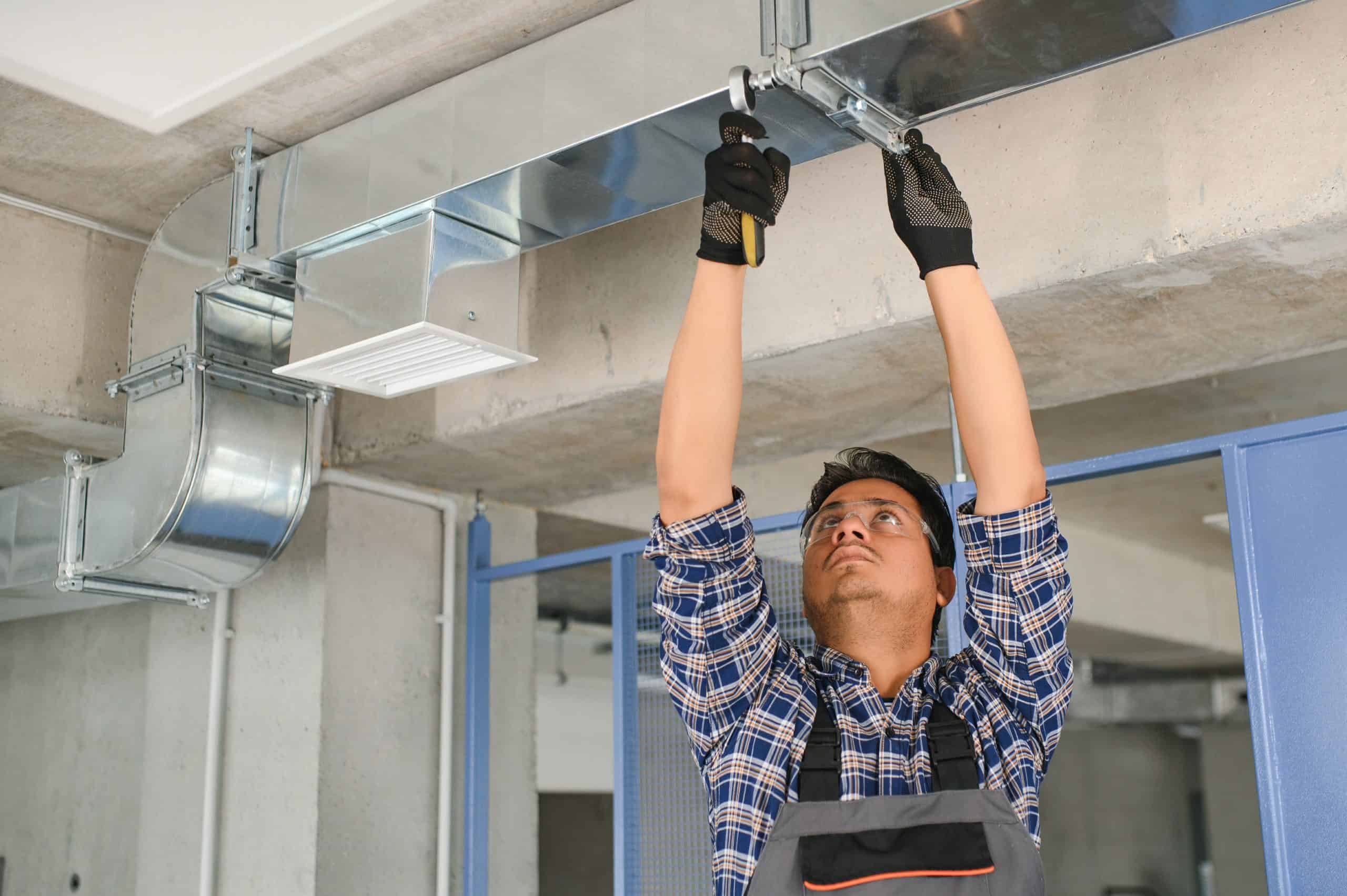 A person wearing a plaid shirt and overalls is standing on a ladder, using tools to install or repair ductwork on a ceiling. The person is wearing black gloves and is in an indoor industrial setting, working on HVAC Cleaning Sacramento to ensure optimal air quality.