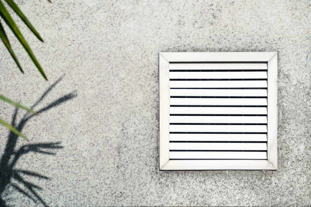 A square ventilation grate with horizontal slats is installed on a textured concrete wall. The shadow of a plant with spiky leaves is cast on the wall to the left of the grate, highlighting areas that may benefit from Residential Duct Cleaning Sacramento for optimal airflow.