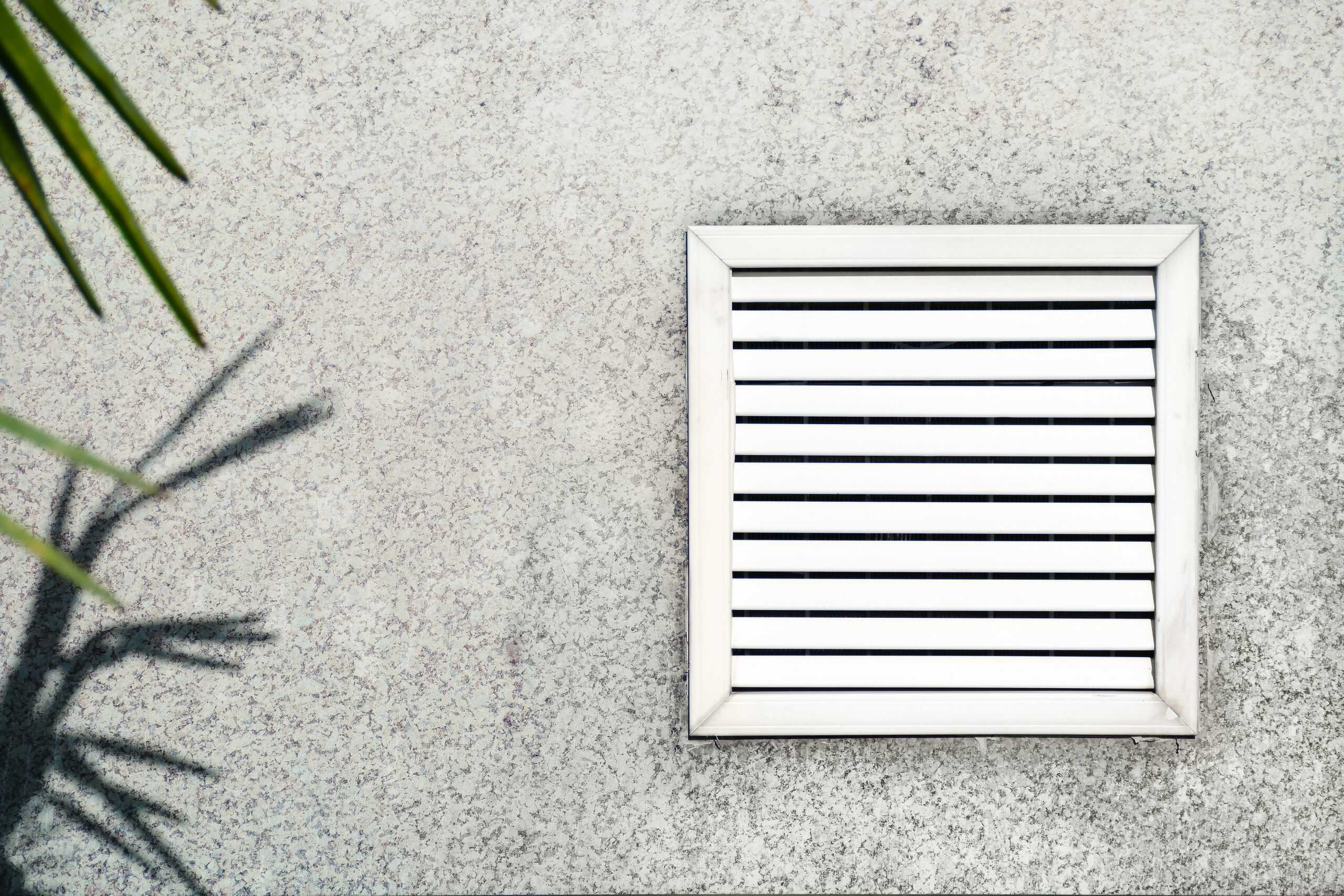 A square ventilation grate with horizontal slats is installed on a textured concrete wall. The shadow of a plant with spiky leaves is cast on the wall to the left of the grate, highlighting areas that may benefit from Residential Duct Cleaning Sacramento for optimal airflow.
