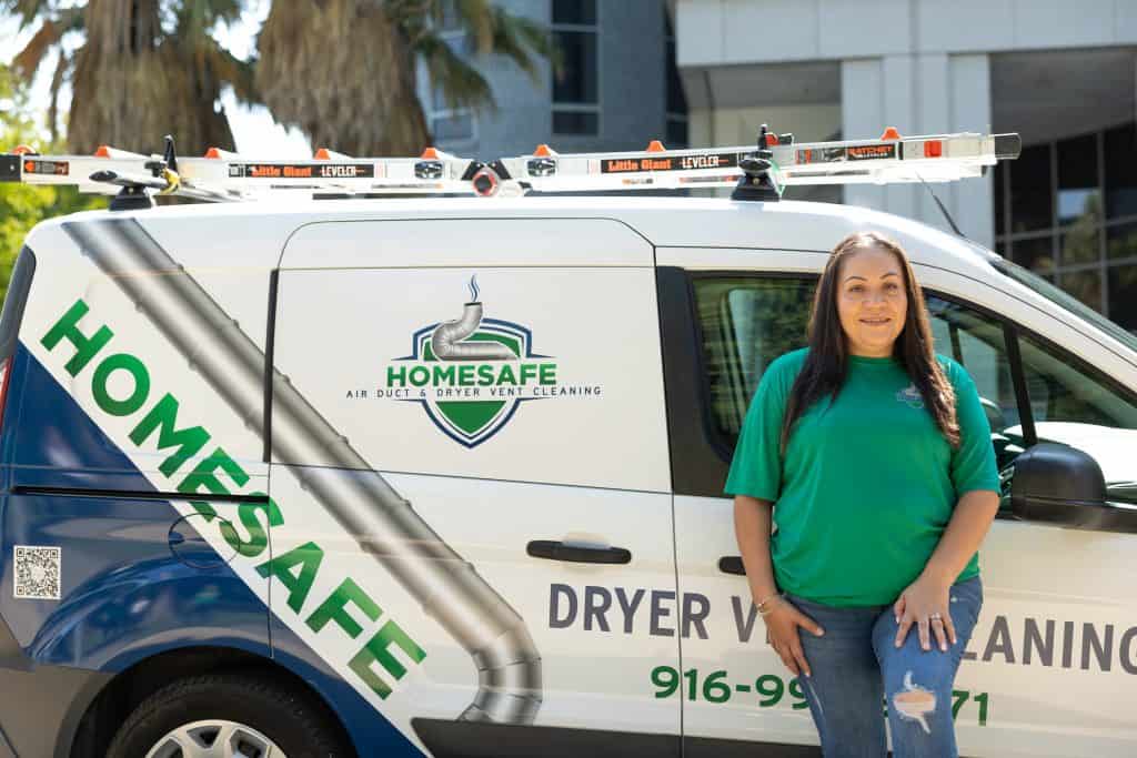 A person in a green shirt smiles, standing beside a white van with "HOMESAFE" and "DRYER VENT CLEANING" signage. The van, equipped for ventilation system cleaning with a ladder on top and contact details, is parked near a building with trees in the background.