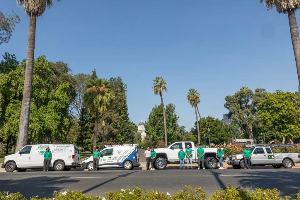 A group of people in green shirts stands in front of several service vehicles, all parked in a row on a road. The vehicles display business logos for HVAC system maintenance. The background features a lush park with palm trees and a distant view of a white-domed building.