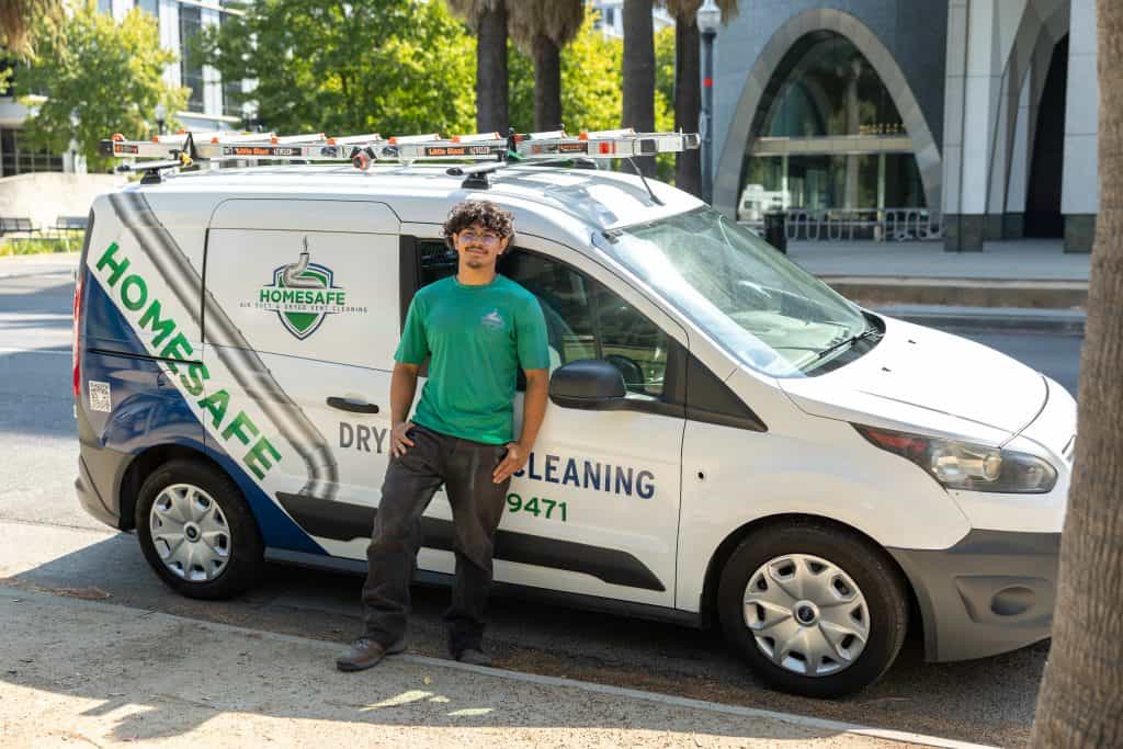 A person wearing a green shirt and dark pants leans against a van labeled "HOMESAFE DRYER CLEANING" with additional contact information. The van, equipped for HVAC cleaning with ladders on the roof rack, is parked on a street adorned with palm trees and modern buildings in the background.