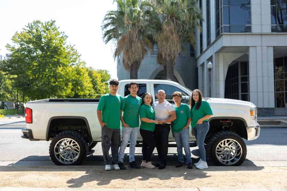 A group of six people wearing green shirts pose in front of a large, white pickup truck. They are standing on a street with trees and a modern building, featuring large windows and palm trees in the background—ready for their next HVAC system maintenance job.