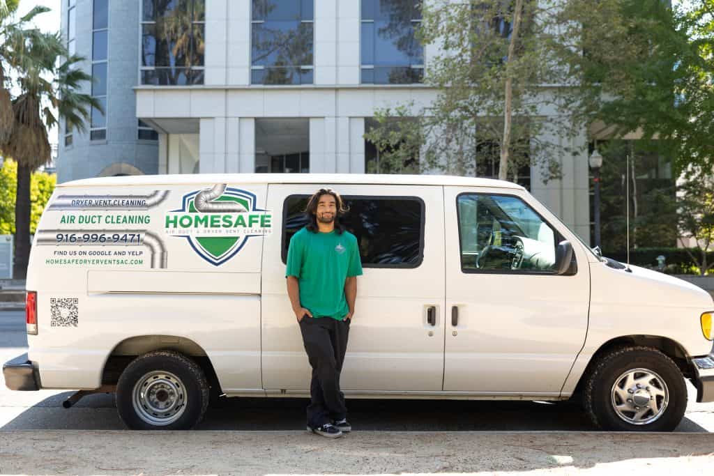 A man wearing a green shirt stands in front of a white van with the "Homesafe Air Duct Cleaning" logo and contact information on its side. The van, parked outdoors with a building and trees in the background, highlights their expertise in ventilation system cleaning.