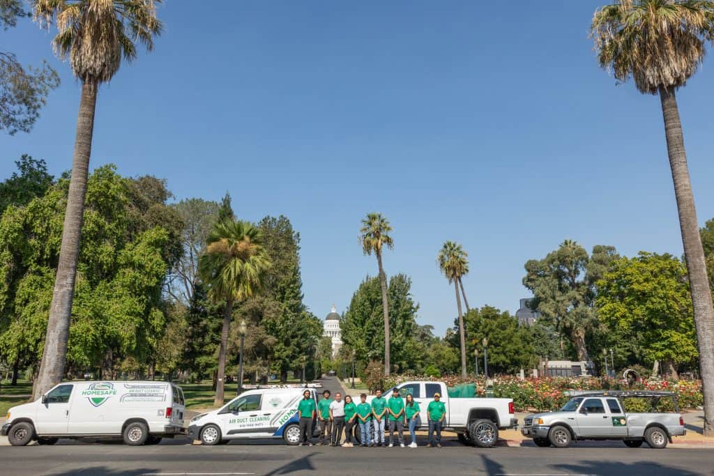 A group of people in matching green shirts stands in front of a row of service vehicles, including trucks and vans labeled with "HVAC System Maintenance," parked along a tree-lined street. The trees and a distant government building with a dome are visible in the background.