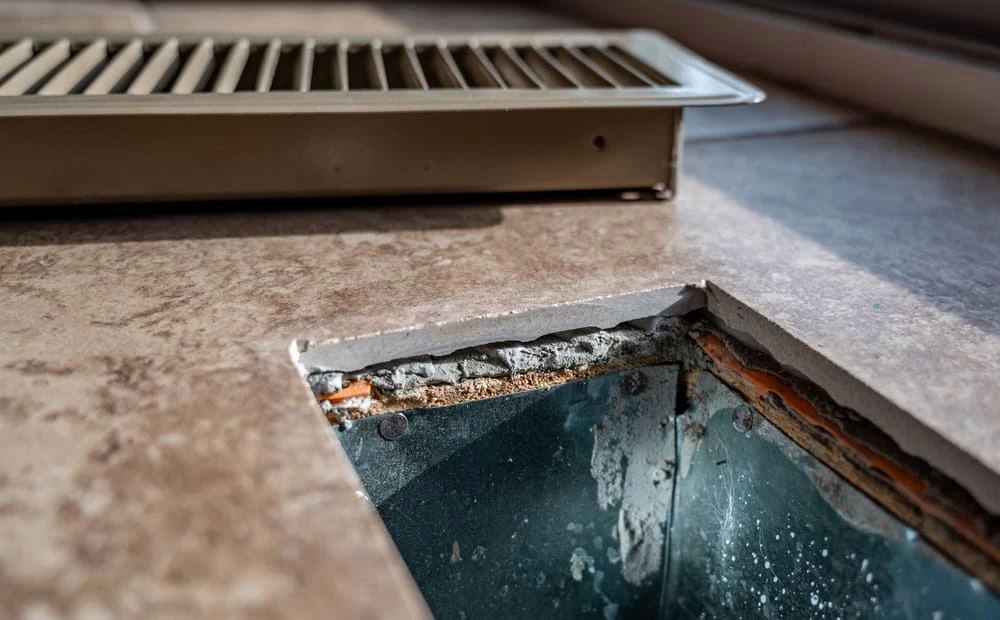 A close-up of a floor vent with the grille removed, revealing the metal ductwork below. The surrounding tile floor shows signs of wear, with chipped edges around the vent opening. The vent cover rests beside the opening, highlighting the need for ventilation system cleaning.
